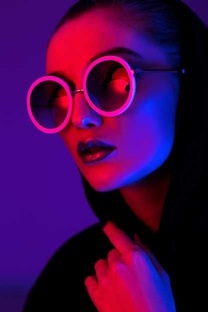 Retrato de moda joven con un suéter negro con capucha y gafas de sol redondas en luz de neón roja y azul en el estudio.