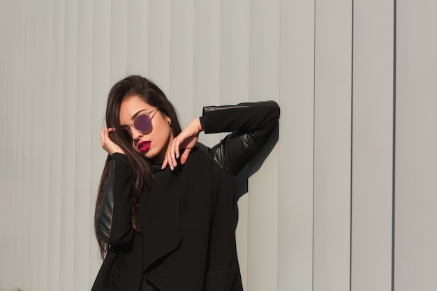 Retrato de moda de modelo de lujo con pelo largo en abrigo de moda y elegantes gafas posando en el edificio