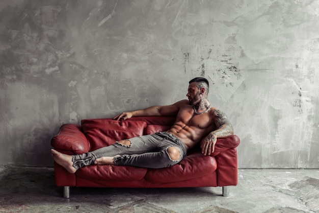 Retrato de moda de modelo masculino desnudo sexy con tatuaje y una barba negra en pose caliente en el sofá de cuero rojo. Interior de la habitación loft con muro de hormigón gris.