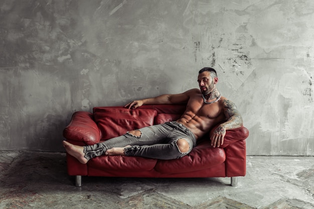 Retrato de moda de modelo masculino desnudo sexy con tatuaje y una barba negra en pose caliente en el sofá de cuero rojo. Interior de la habitación loft con muro de hormigón gris.