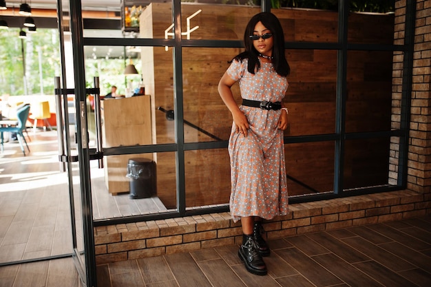 Retrato de moda de mujer afroamericana con vestido largo, botas y gafas de sol en el interior.