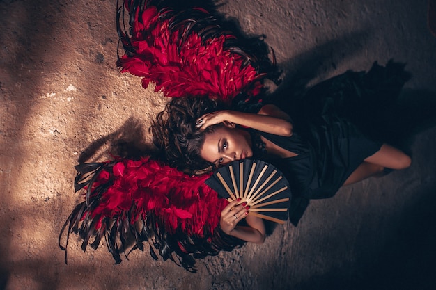 Retrato de moda de una mujer apasionada con un vestido negro con alas de ángel rojo y con abanico negro en la mano. Concepto de moda de belleza oscura.