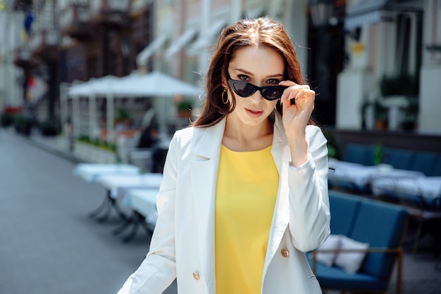 Retrato de moda de mujer atractiva en chaqueta blanca con pelo largo. Encantadora dama de gafas de sol mirando a la cámara. Feliz chica con estilo en la calle de la ciudad.