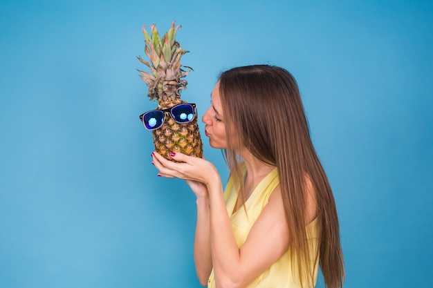 Retrato de moda mujer besa piña en gafas de sol sobre fondo azul.