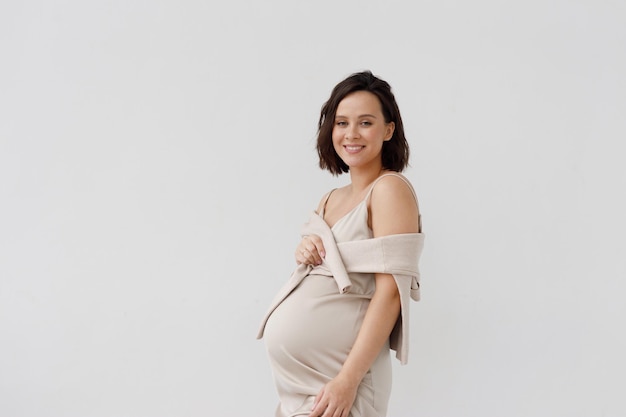 Retrato de moda de una mujer embarazada feliz con un vestido elegante. Una niña contra una pared blanca con un espacio vacío.