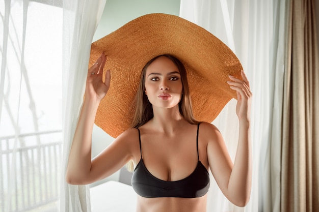 Retrato de moda: mujer encantadora y atractiva con maquillaje posando en un gran sombrero de paja frente a la ventana. Vacaciones tropicales