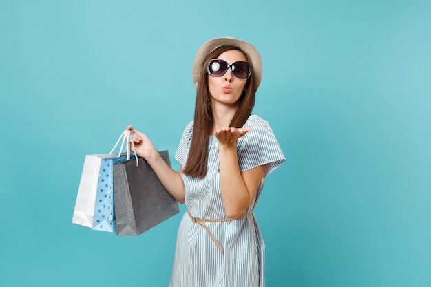 Retrato de moda mujer feliz atractiva en vestido de verano, sombrero de paja, gafas de sol con bolsas de paquetes con compras después de ir de compras aislado sobre fondo azul pastel. Copie el espacio para publicidad.