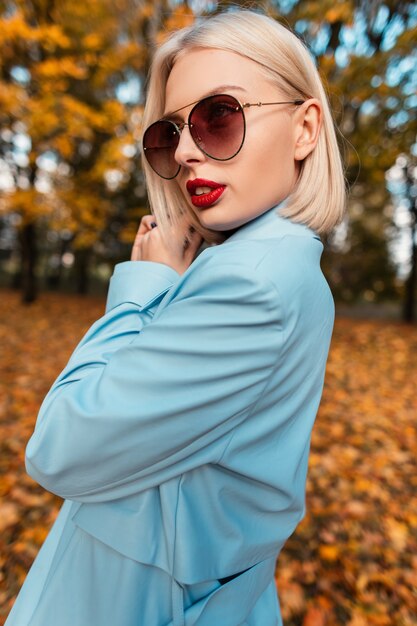 Retrato de moda de una mujer hermosa con un corte de pelo corto y labios rojos con ropa de moda con un abrigo azul y gafas de sol Ð¼ÑˆÑ‚ÐµÑ „Ð¿Ñƒ en la naturaleza en un parque otoñal con follaje amarillo