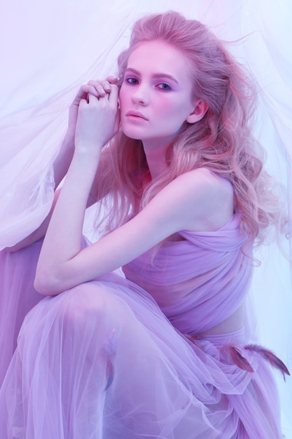 Retrato de moda de mujer hermosa joven en vestido violeta esponjoso. Cabello rubio rizado, maquillaje, vestido con plumas, colores suaves, fotografía artística