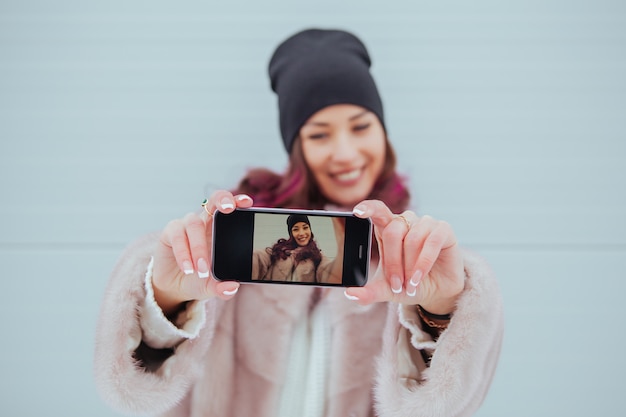 Retrato de moda de mujer hipster muy sonriente en abrigo de visón y smartphone contra la colorida pared gris. Haciendo selfie Ropa casual. Pelos de colores