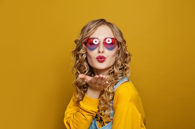 Retrato de moda de una mujer joven bonita con el pelo largo rubio y rizado soplando un beso en amarillo brillante