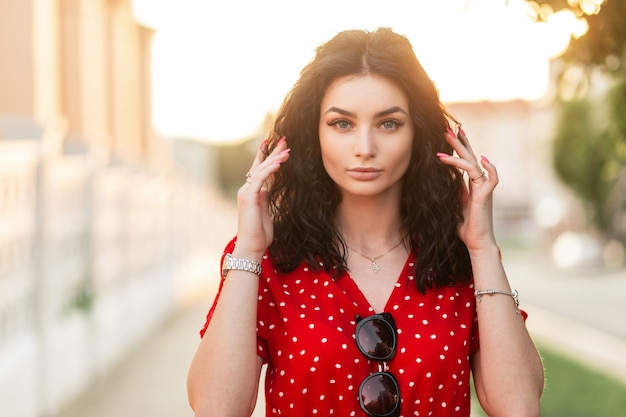 Retrato de moda de una mujer joven y bonita con el pelo rizado en un vestido rojo vintage con gafas de sol y reloj de joyería en la calle al atardecer