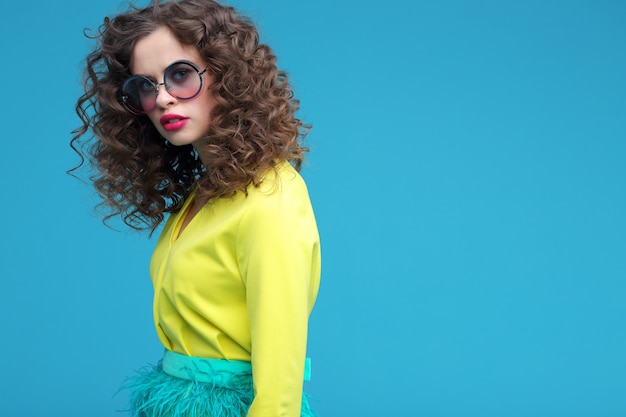 Retrato de moda de mujer joven hermosa con un vestido amarillo de verano. Cabello rizado, gafas de sol, fondo azul.