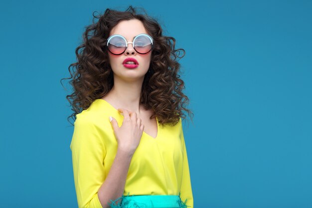 Retrato de moda de mujer joven hermosa con un vestido amarillo de verano. Cabello rizado, gafas de sol, fondo azul.