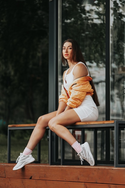 Retrato de moda de mujer de moda joven vestida con vestido blanco, chaqueta y zapatillas de deporte sentado en el banco contra el café de la ciudad, moda callejera de verano. retrato de mujer alegre