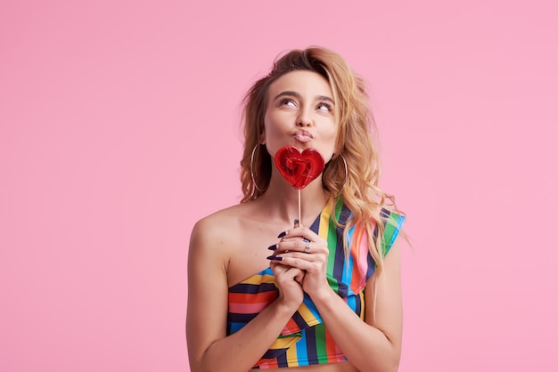 Retrato de moda de mujer sonriente joven muy dulce divirtiéndose con piruleta en rosa. Muestra un dedo al lado derecho. Copiar espacio para texto