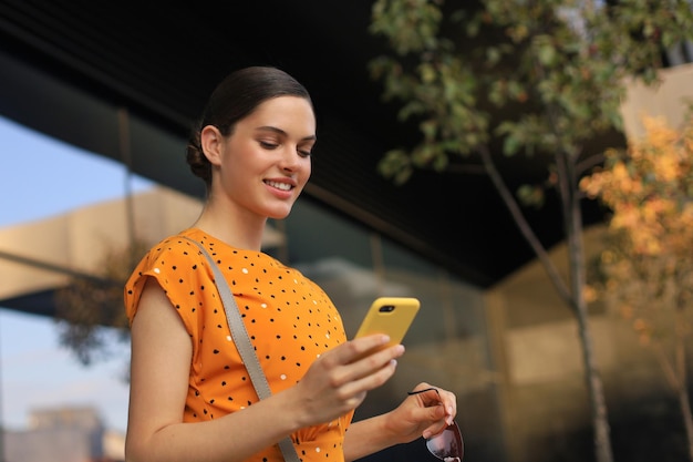 Retrato moda mulher de vestido amarelo andando na rua e segurando o smartphone na mão