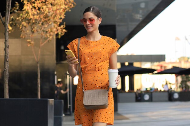 Retrato moda mulher de vestido amarelo em óculos de sol andando na rua e segurando o smartphone na mão