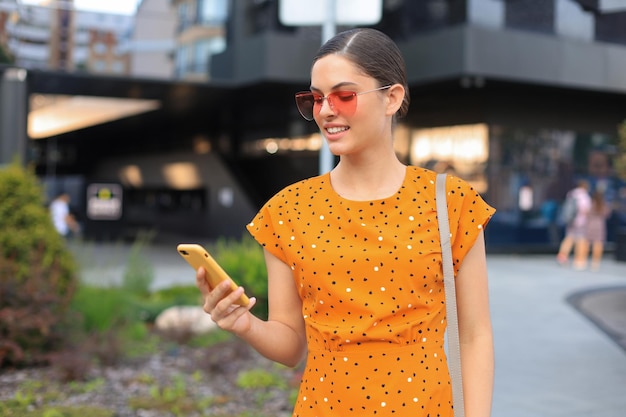 Retrato moda mulher de vestido amarelo em óculos de sol andando na rua e segurando o smartphone na mão