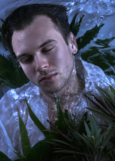 Retrato de moda de primer plano de hombre caucásico guapo sin afeitar bajo el agua en sexy camisa blanca mojada ojos cerrados cara fuerte, fondo de plantas