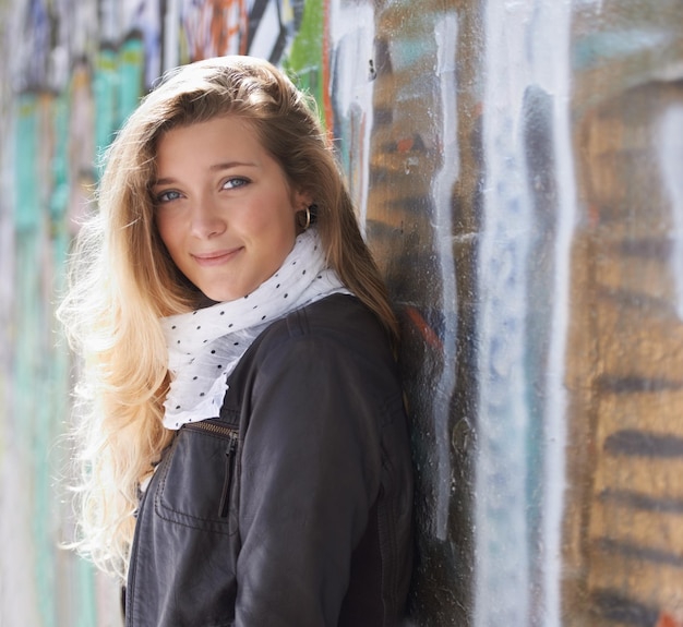 Retrato de moda urbana y mujer en la pared con graffiti de sonrisa y de pie en el arte callejero al aire libre Felicidad juventud y cara feliz de la modelo gen z en la ciudad con ropa de calle estilo invernal y sol