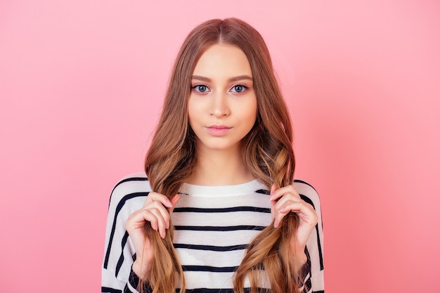 Retrato de un modelo elegante de mujer de pelo largo adolescente joven y atractivo en un suéter de rayas en estudio sobre un fondo rosa. concepto de cabello sano, fuerte y grueso