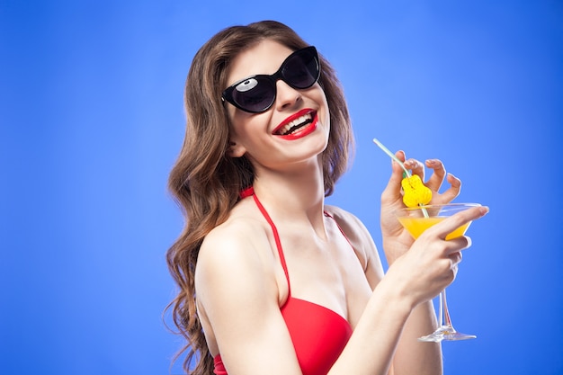 Retrato de modelo morena feliz con cóctel y gafas de sol sonriendo a la cámara.