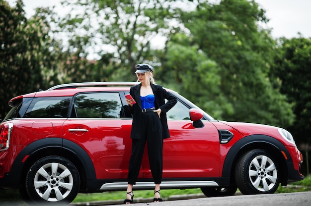 Retrato de modelo de mujer de moda sexy rubia hermosa en gorra y en todo negro con maquillaje brillante cerca de coche rojo de la ciudad, con teléfono móvil.