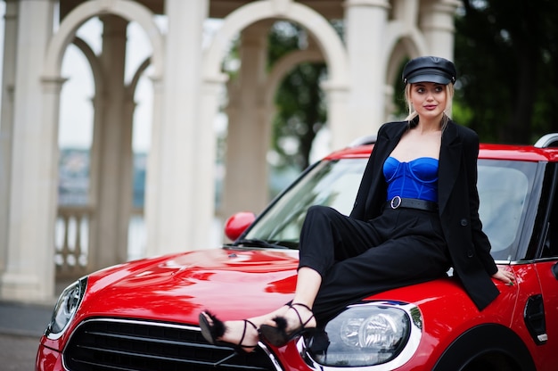 Retrato de modelo de mujer de moda sexy rubia hermosa en gorra y en todo negro con maquillaje brillante sentarse en el capó del coche rojo de la ciudad.