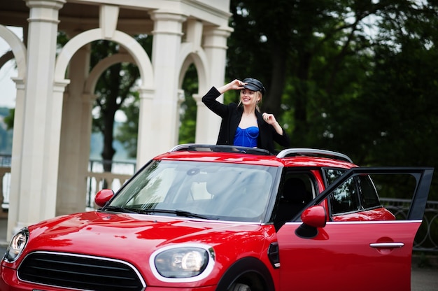 Retrato de modelo de mujer de moda sexy rubia hermosa en gorra y en todo negro con maquillaje brillante en techo corredizo abierto de coche rojo de la ciudad.