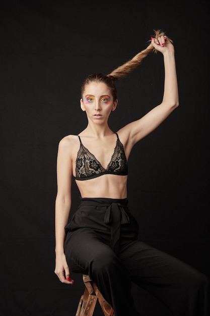 Retrato de una modelo rubia con sujetador y pantalones negros sosteniendo su cabello, sentada en una escalera de madera. En estudio sobre fondo negro.