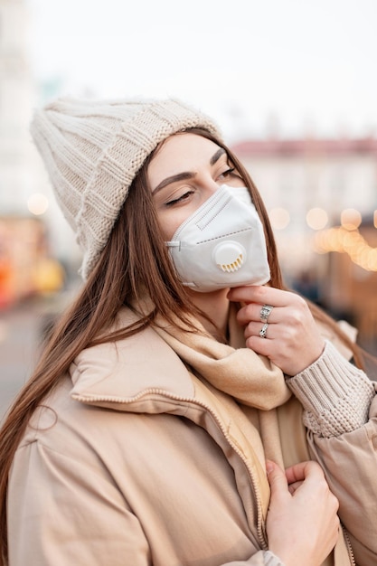 Retrato moderno de uma bela jovem com uma máscara de proteção médica em roupas de inverno da moda com um chapéu de malha, suéter e jaqueta anda na rua