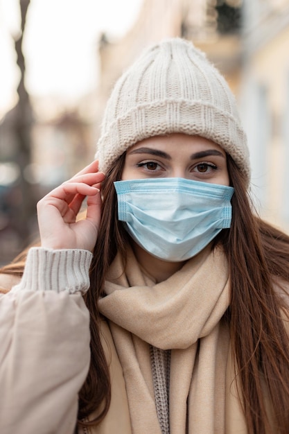 Retrato moderno de una joven mujer bonita con una máscara protectora médica en ropa de invierno de moda con un sombrero de punto, chaqueta y bufanda camina por la ciudad y mira la cámara. estilo COVID-19