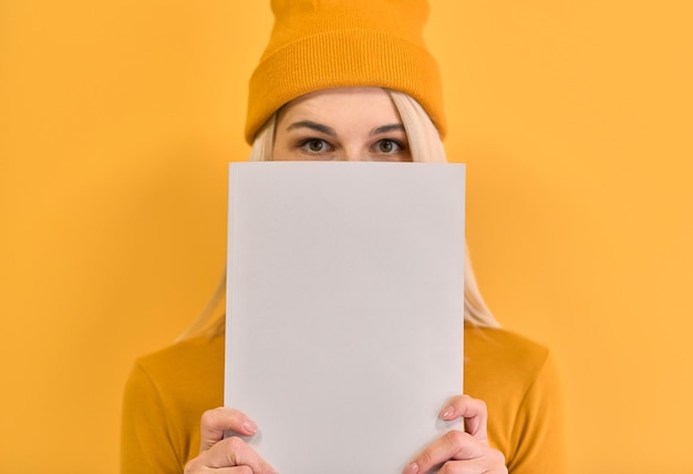 Retrato monocromático de primer plano interior de una mujer alegre que cubre la cara con un libro blanco tiene una expresión alegre espacio en blanco para su publicidad o texto promocional aislado en el fondo amarillo