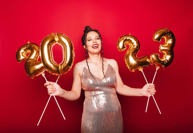 Retrato de una morena sobre un fondo rojo sosteniendo globos dorados en forma de 2023 próximo año nuevo espacio de Navidad para texto