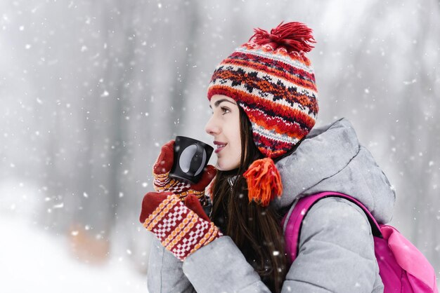 Retrato de una morena turista con chaqueta gris, sombrero y mitones inhala el aroma del té caliente en el bosque frío de invierno