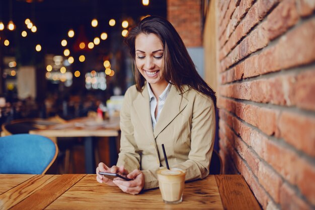 El retrato de la morenita caucásica hermosa vistió la sentada casual elegante en café y el uso del teléfono inteligente para enviar el mensaje. En la mesa de madera hay un delicioso café recién hecho.