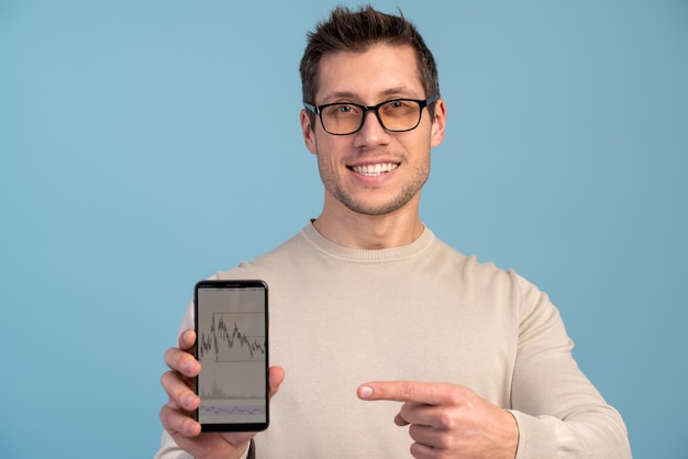 Retrato de un moreno satisfecho con barba apuntando al celular y sonriendo a la cámara, recomendando un gadget o una aplicación móvil. Disparo de estudio interior aislado sobre fondo azul.