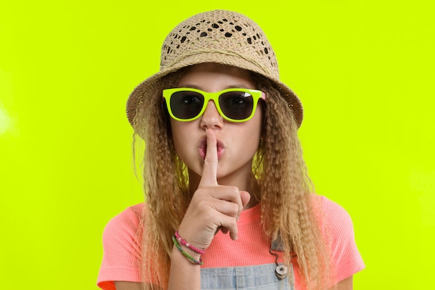 Retrato de muchacha adolescente en sombrero de gafas de sol con el dedo índice cerca de los labios, mostrando el signo de silencio