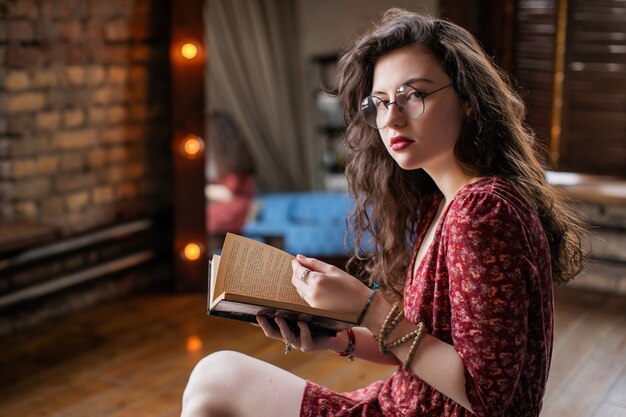 Retrato de muchacha atractiva estudiante con el pelo rizado, en vestido rojo corto y gafas redondas. Mujer joven que mira in camera mientras que se sienta en el apartamento del desván y lee el libro en sus manos.