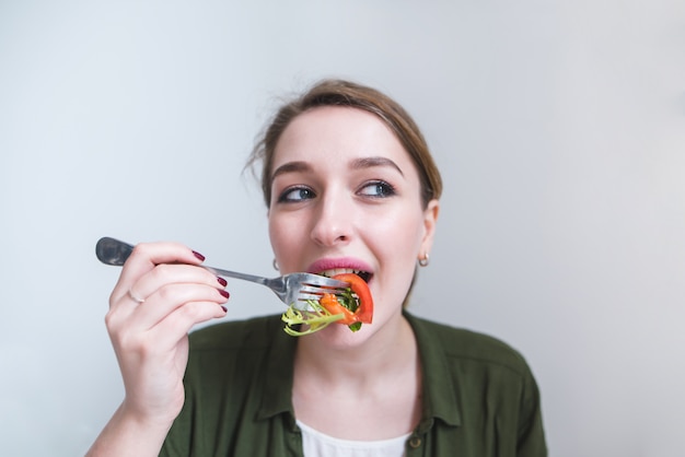 Retrato de la muchacha bonita que come la ensalada con la bifurcación en fondo gris. hermosa mujer come comida saludable.