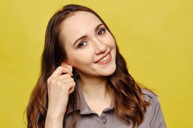 El retrato de una muchacha caucásica hermosa toca el oído en un vestido gris en un fondo amarillo.