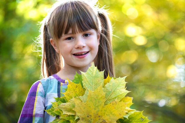 El retrato de la muchacha feliz del niño bonito que sostiene un manojo de árbol caido se va en bosque del otoño. Niño femenino positivo disfrutando de un día cálido en el parque de otoño.