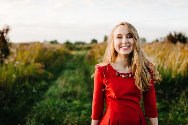 El retrato de una muchacha hermosa se coloca en otoño en un vestido rojo contra el campo en la naturaleza. mitad superior. mira hacia adelante. De cerca. El viento disipó el cabello.