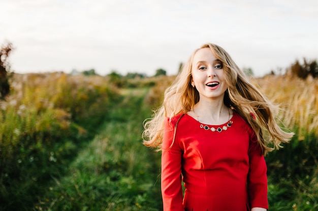 El retrato de una muchacha hermosa se coloca en otoño en un vestido rojo contra la perspectiva del campo en la naturaleza.