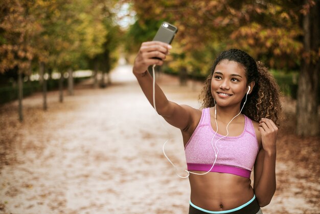 Retrato de una muchacha joven y atractiva de los deportes que toma un selfie en su teléfono y música que escucha en el parque.