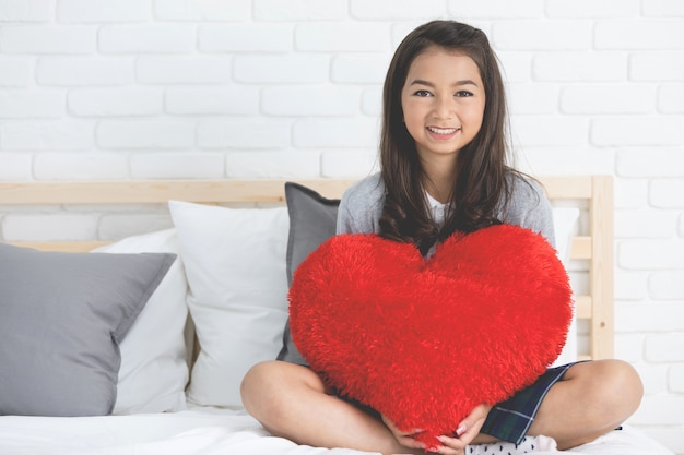 El retrato de la muchacha linda asiática se sienta en la cama con el corazón rojo grande de la forma de la almohada.