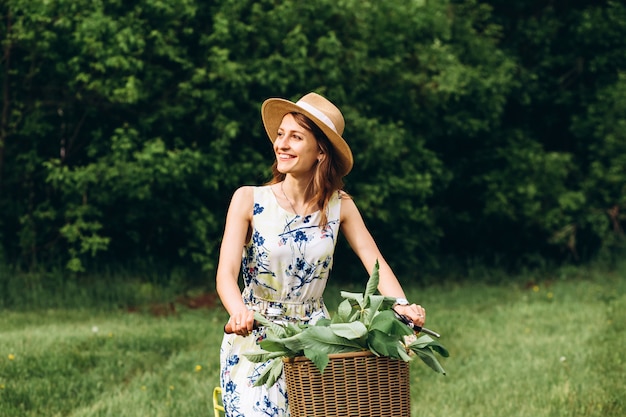 Retrato de muchacha linda con el pelo rubio rizado en sombrero conduciendo una bicicleta. Personas activas Al aire libre. Hermosa chica en bicicleta en el campo que huele a flores, estilo de vida de verano. primer retrato