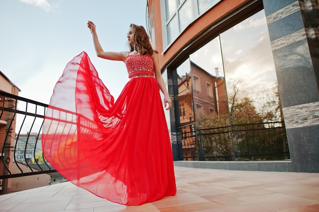 El retrato de la muchacha de moda en el vestido de noche rojo presentó la ventana del espejo del fondo del edificio moderno en el balcón de la terraza. Vestido que sopla en el aire