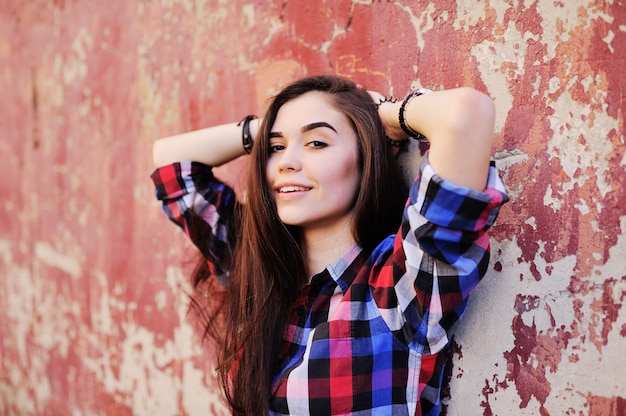 Retrato de la muchacha morena joven linda en camisa de tela escocesa en fondo rojo de la pared del grunge.